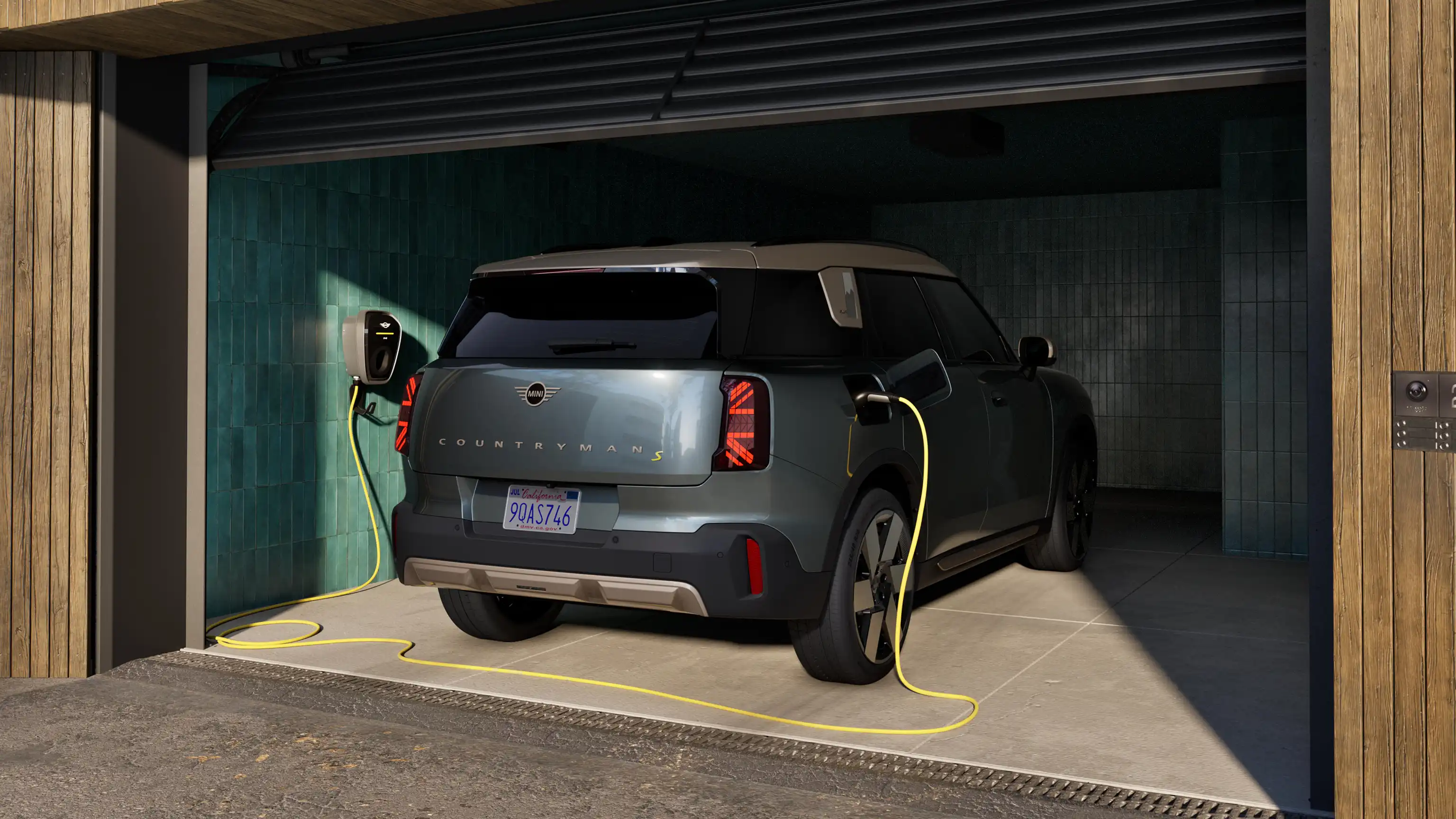 Rear view of charging all-electric MINI Countryman in Smokey Green standing in open garage connected with charging cable.