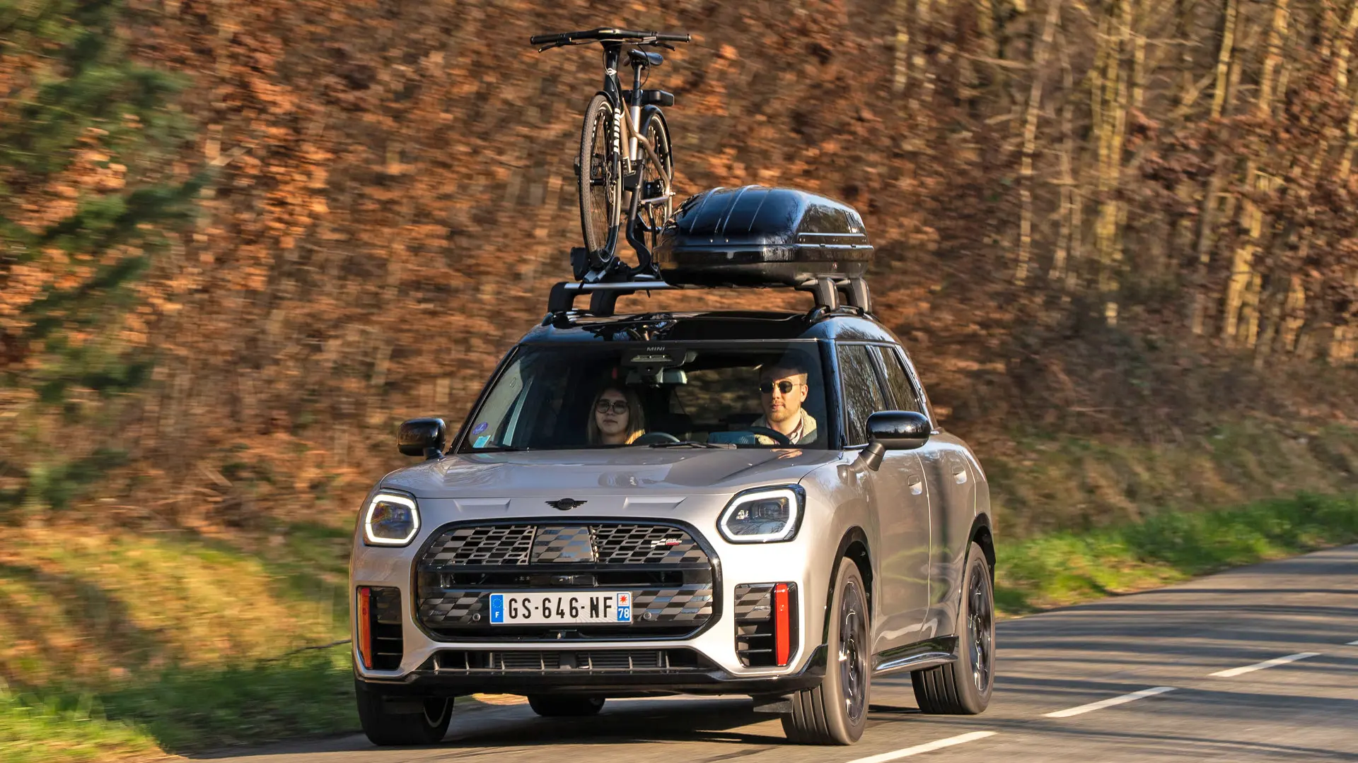 MINI Countryman Cooper in Silver on a mountain road with MINI Roof Rack, MINI Roof Box, MINI Roof Cycle Carrier with one bike mounted plus MINI John Cooper Works winglet – front view. 