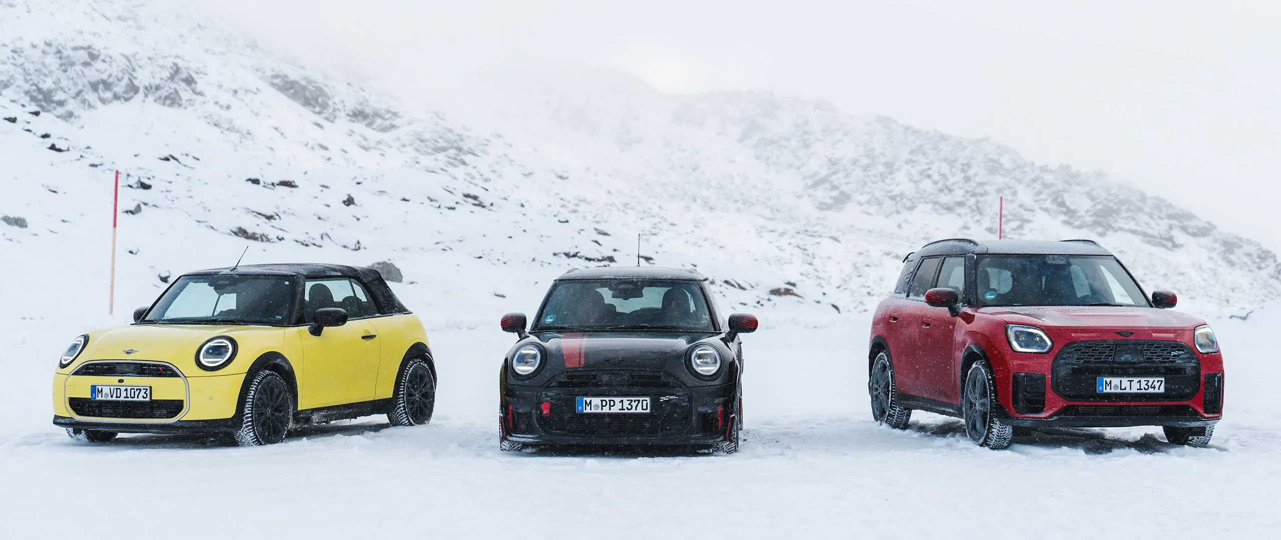 MINI Cooper Convertible in Sunny Side Yellow, MINI John Cooper Works in Midnight Black II, and MINI Countryman Cooper in Chilli Red – stationary on snowy mountain road, front view.